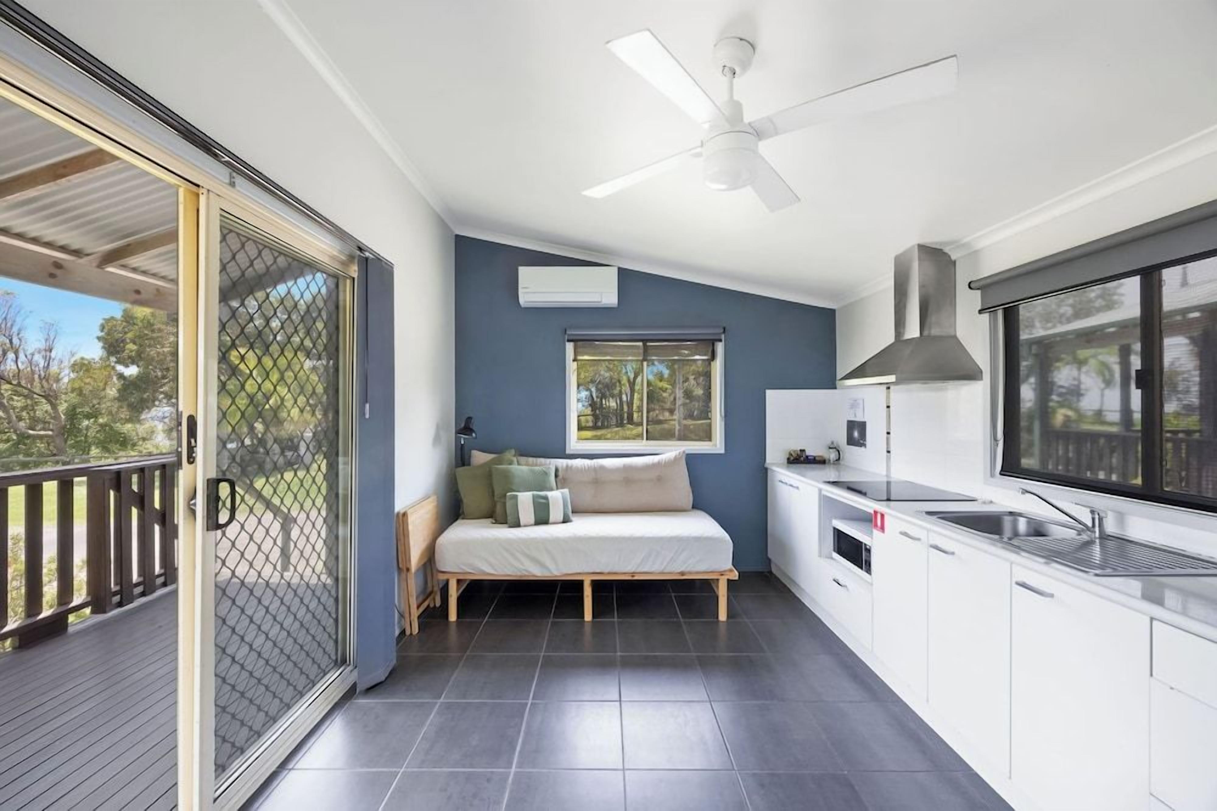 A compact living space with a balcony, a bed, a kitchenette, and a ceiling fan.