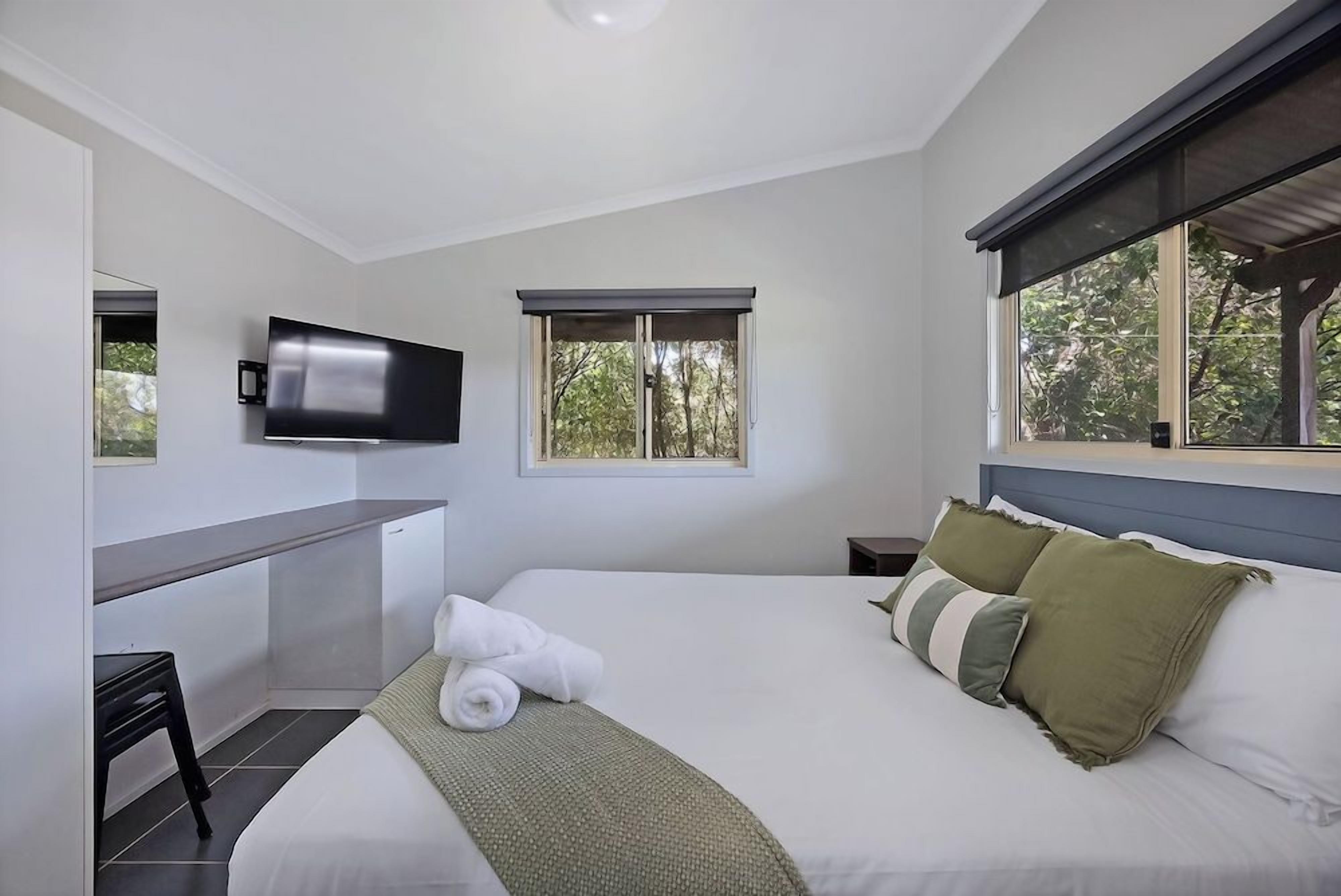 A bedroom with a bed, a TV mounted on the wall, a window, and a small desk area.