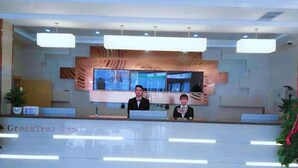 A reception desk with a large screen displaying a news channel, two staff members behind the counter, and a sign reading 'GreenTree Inn'. - Greentree Inn Linxia Autonomous Prefecture Linxia Oxford River Avenue Express Hotel (Linxia)