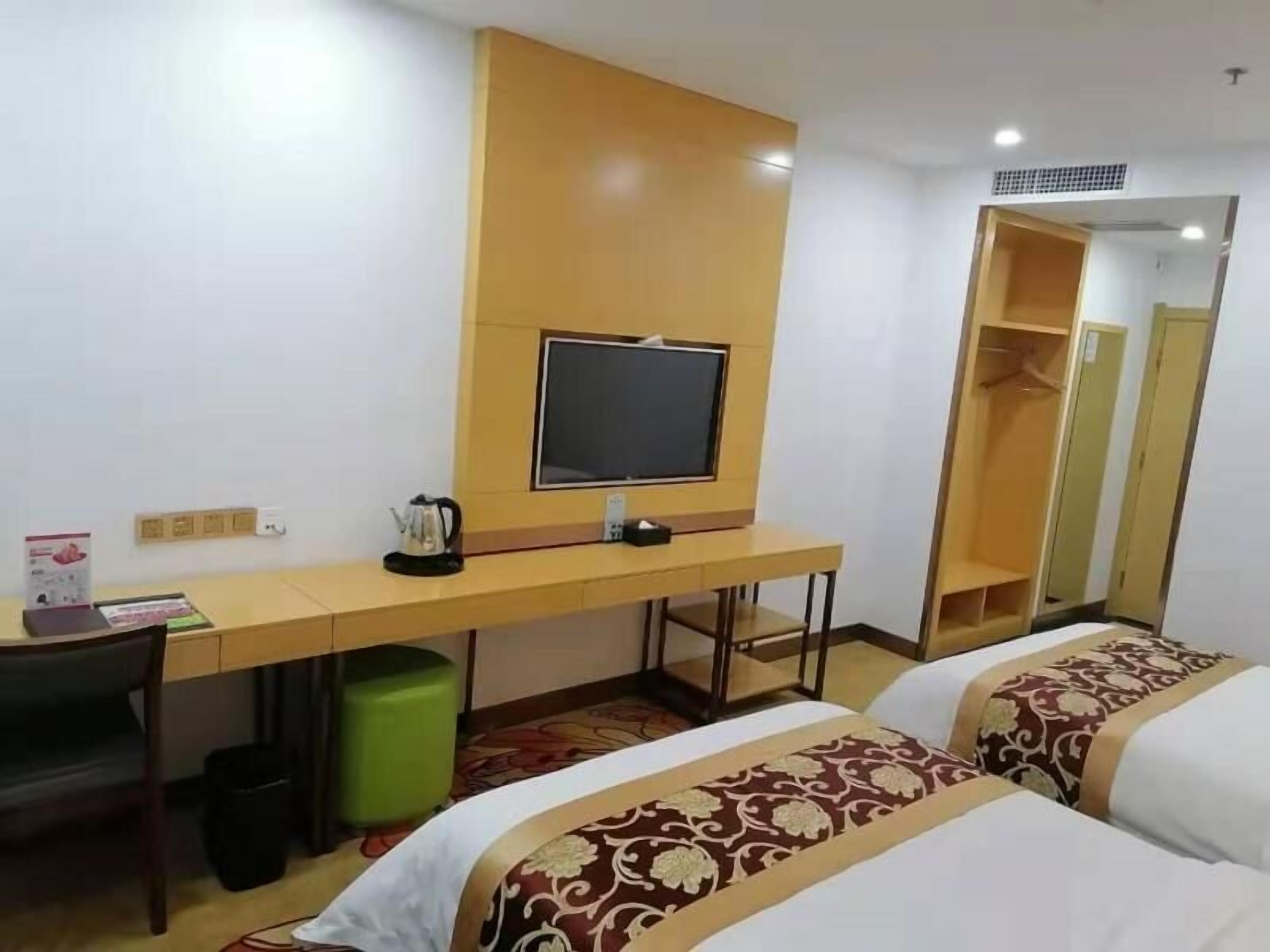 A hotel room with a desk, a television, a bed with patterned bedding, a green trash bin, and a wardrobe.