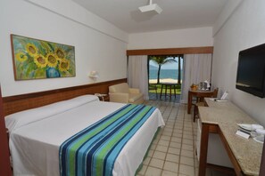 A hotel room with a bed, a desk, a chair, a TV, a painting, and a view of the beach. - Portobello Hotel Resort And Safari (Mangaratiba)