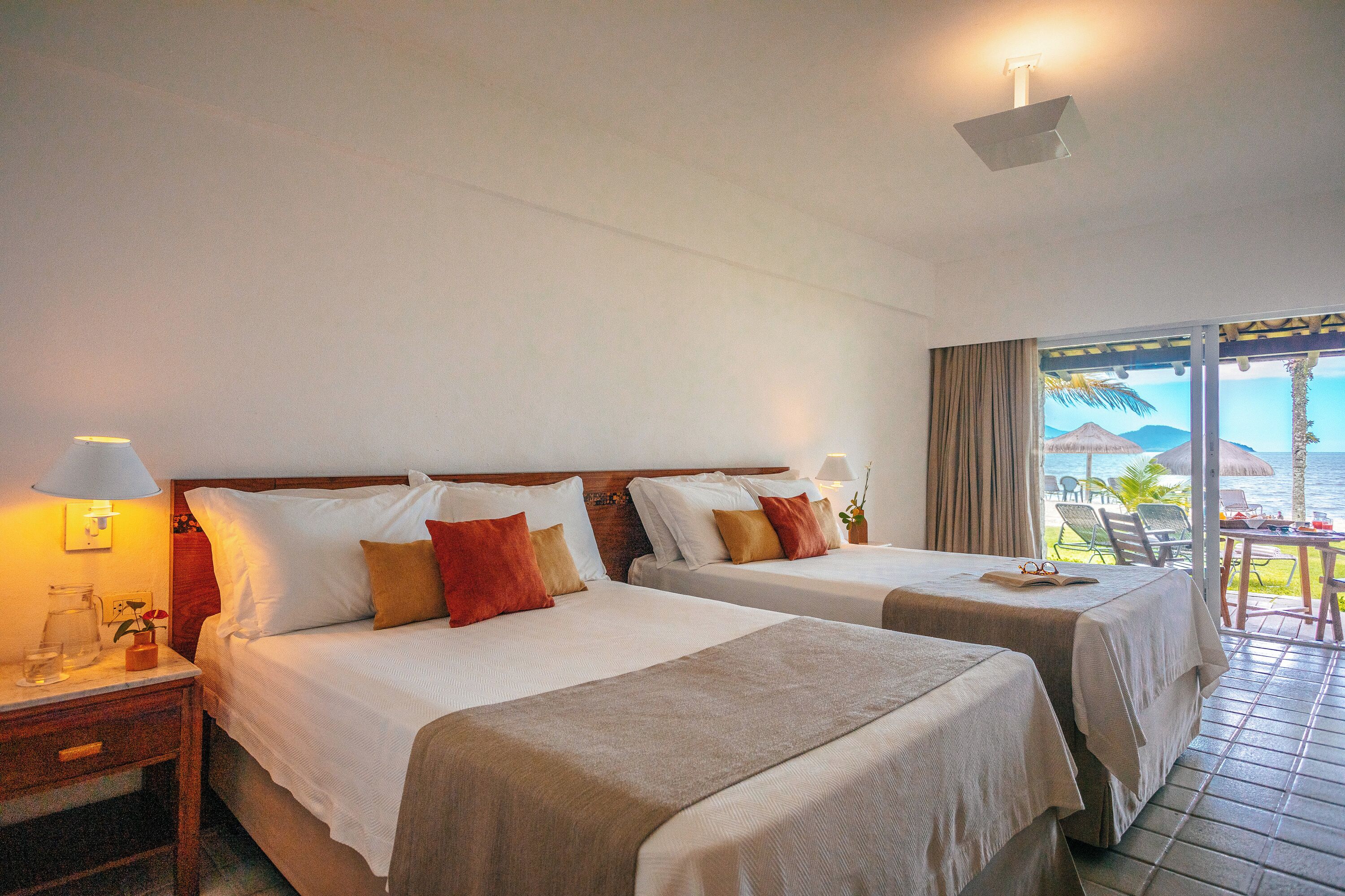 Quarto de hotel com duas camas, uma mesa de cabeceira, uma mesa com abajur e vista para o oceano através de uma porta de correr.