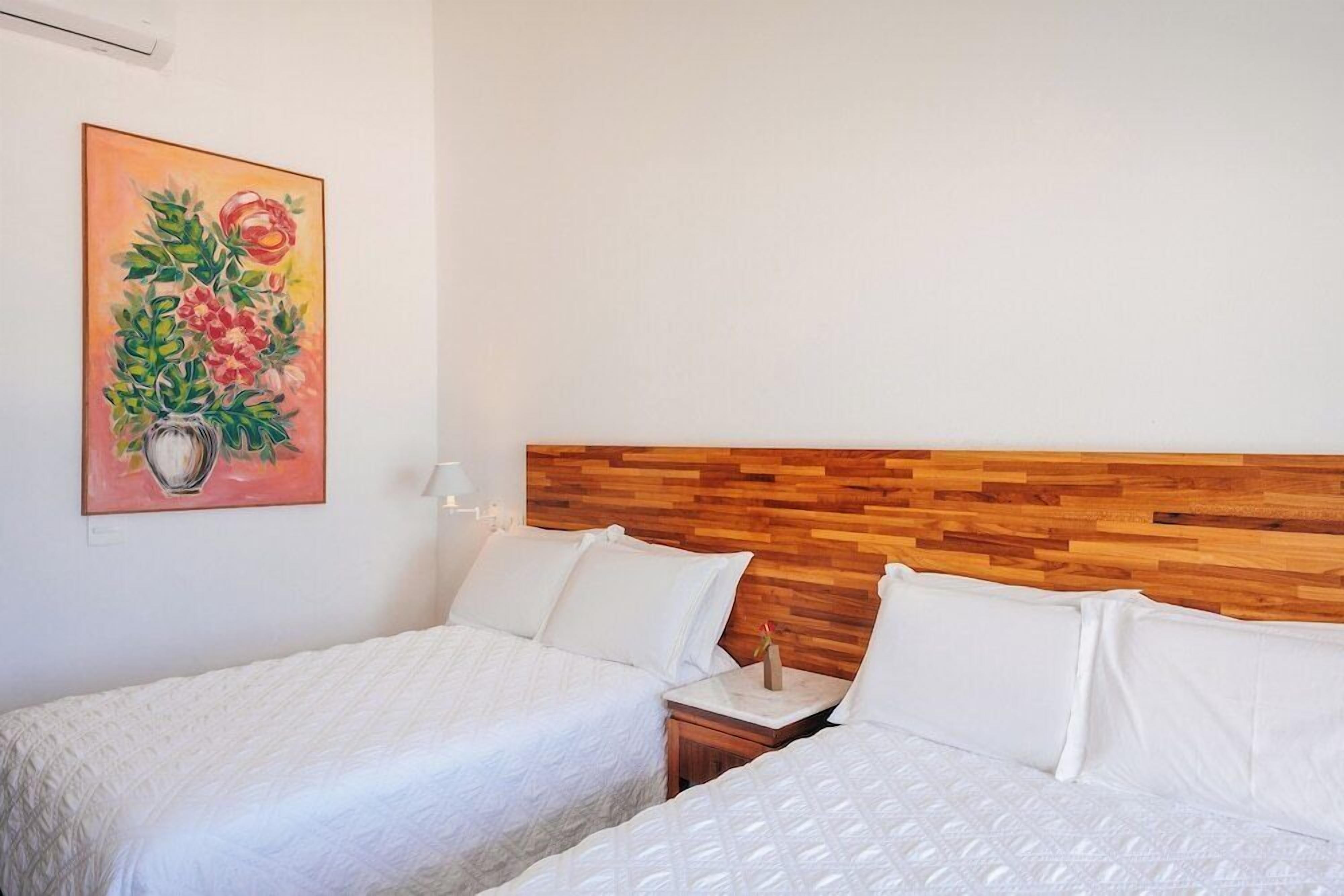 Quarto de hotel com duas camas, uma cabeceira de madeira, um quadro de flores e uma mesa de cabeceira com abajur.