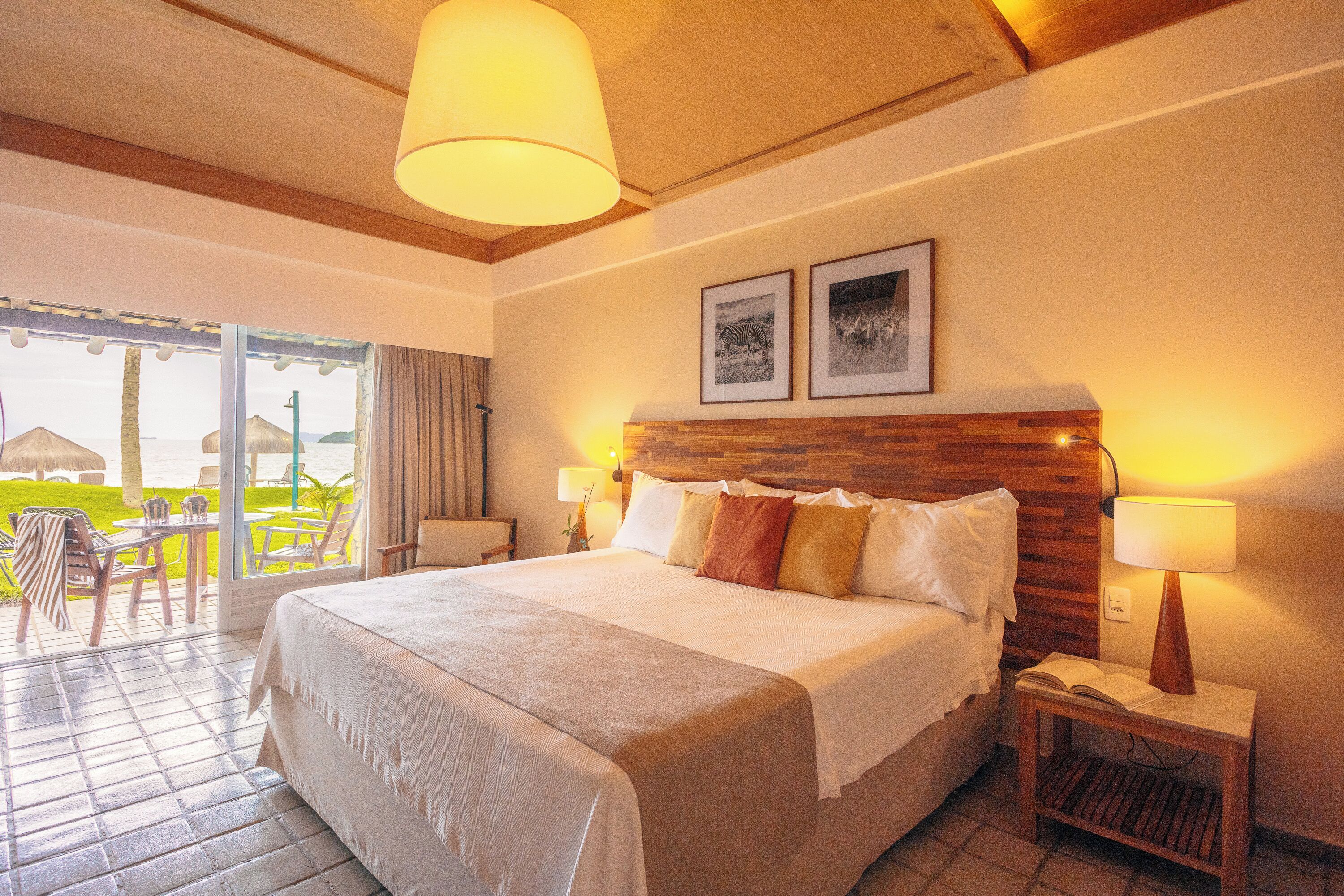 A hotel room with a large bed, wooden headboard, two bedside lamps, and a view of the outdoor area with chairs and umbrellas.
