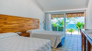 Quarto de hotel com duas camas, cabeceira de madeira, varanda com vista para o mar e área de refeições.
