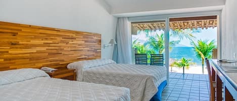 Habitación de hotel con dos camas, cabecera de madera, balcón con vista al mar y zona de comedor.
