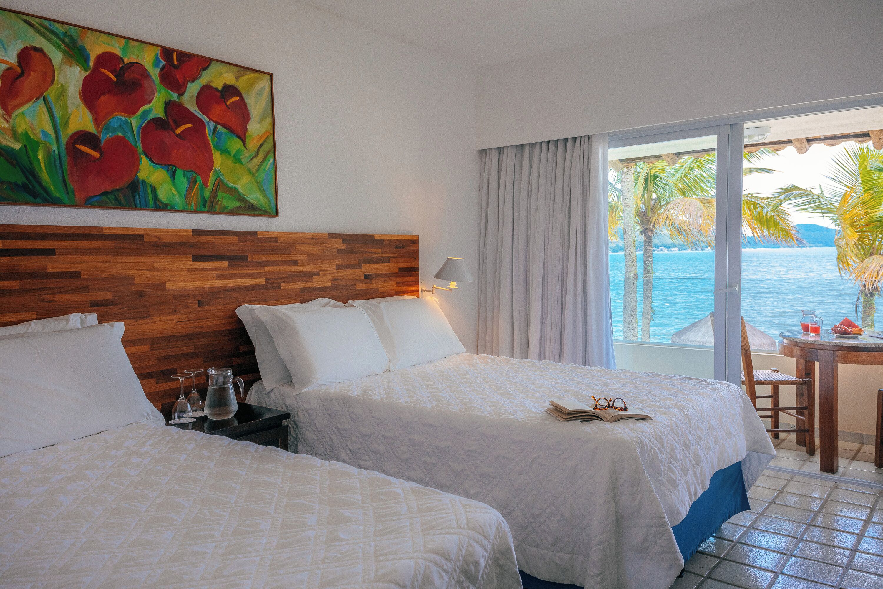 Quarto de hotel com duas camas, cabeceira de madeira, um quadro, varanda com mesa e cadeiras, e vista para o mar.