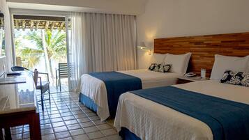 Habitación de hotel con dos camas, un escritorio, una silla, cabecera de madera y vista a palmeras.