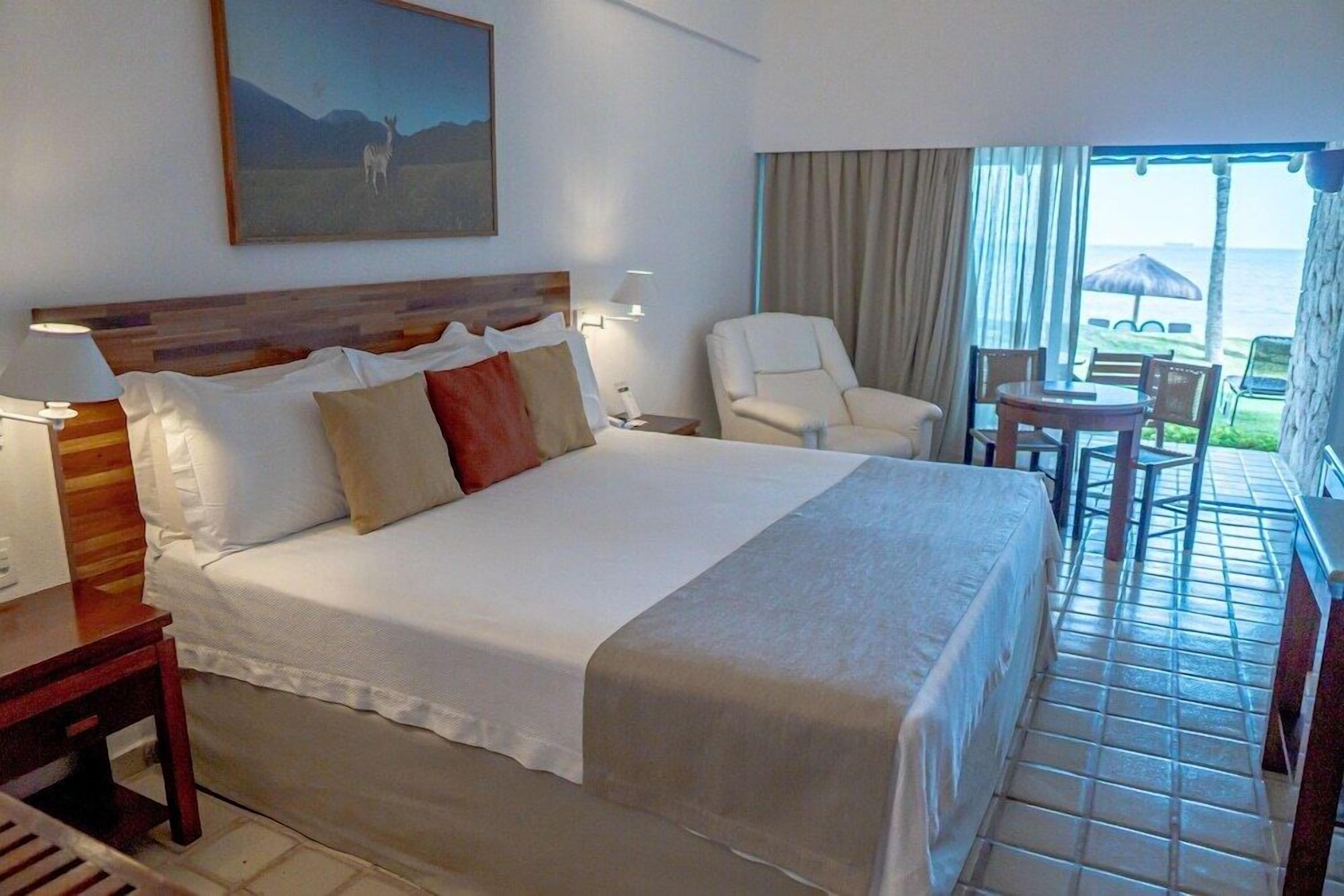 Quarto de hotel com uma cama grande, uma cadeira, uma mesinha, um quadro na parede e vista para o oceano.