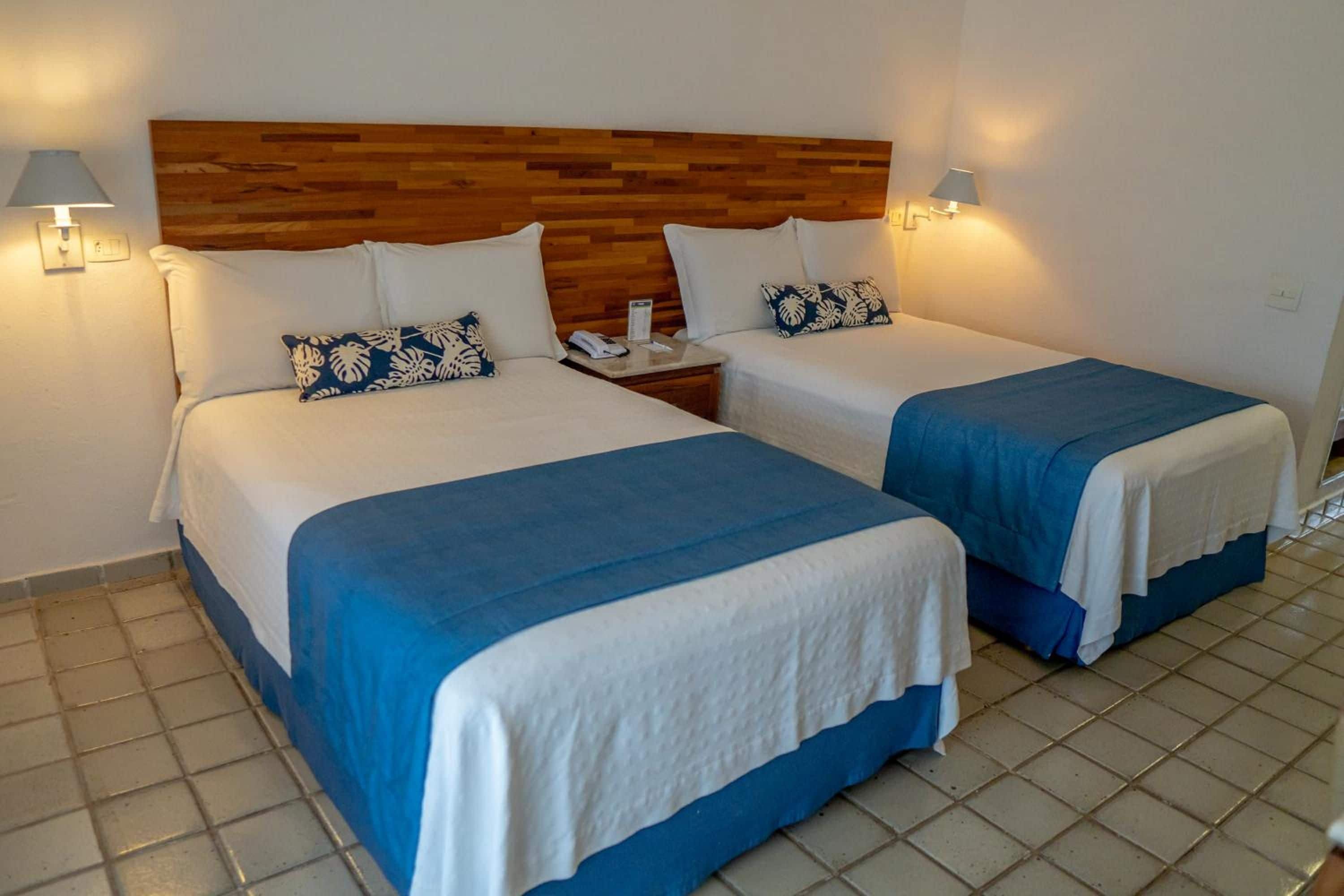 Quarto de hotel com duas camas, cada uma com cabeceira de madeira, roupa de cama branca e detalhes em azul. Há luminárias laterais e uma mesa de cabeceira com telefone e papéis.