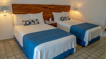 Quarto de hotel com duas camas, cada uma com cabeceira de madeira, roupa de cama branca e detalhes em azul. Há luminárias laterais e uma mesa de cabeceira com telefone e papéis.