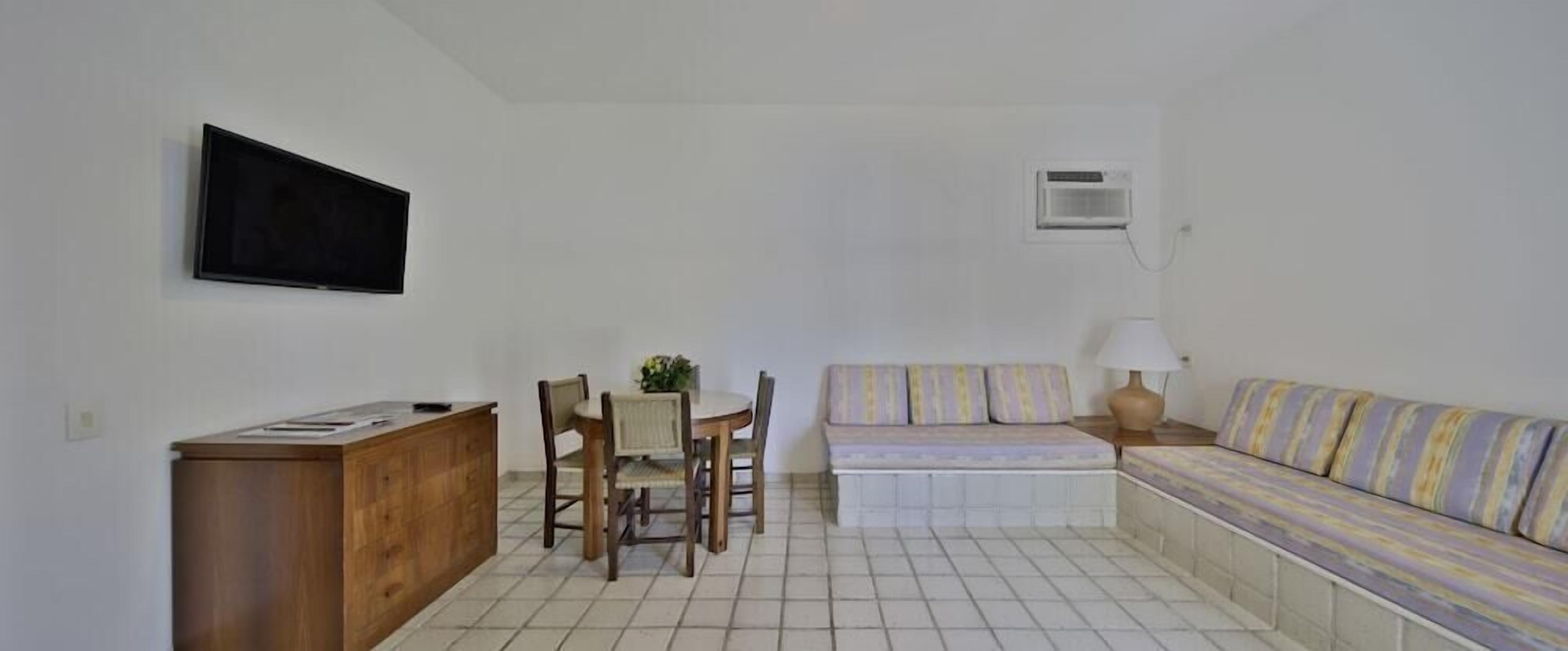 Quarto com escrivaninha de madeira, televisão, sofá, mesa pequena com cadeiras e luminária.