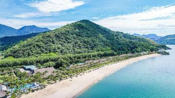 Um resort costeiro com praia, área de piscina e montanhas ao fundo.