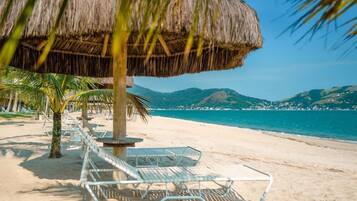 Een strand met rieten parasols, ligstoelen en palmbomen.