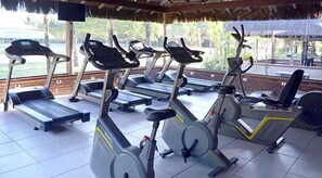 Espaço de ginástica ao ar livre coberto, com bicicletas ergométricas e esteiras.