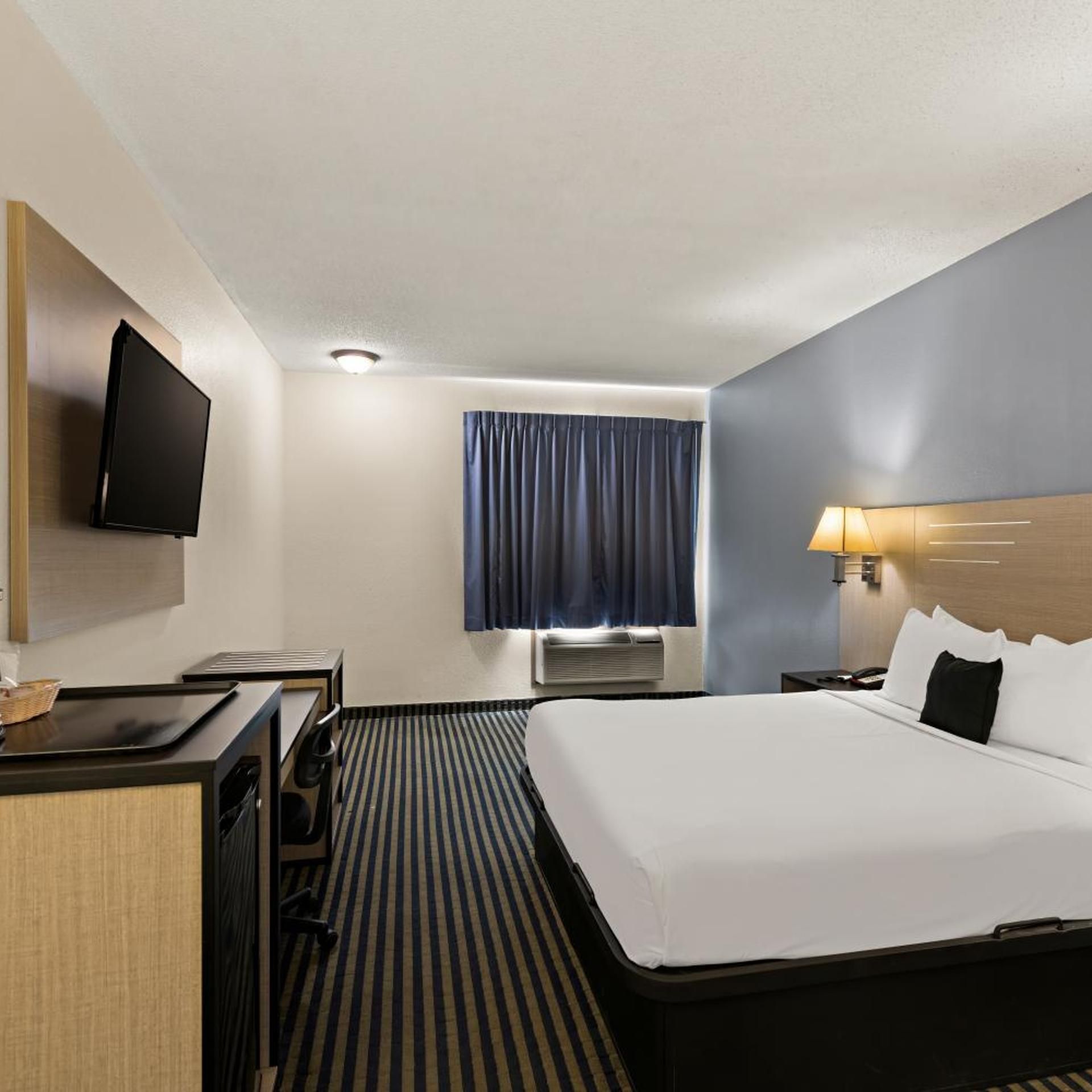 A hotel room with a bed, a desk, a television, a window with curtains, and an air conditioning unit.