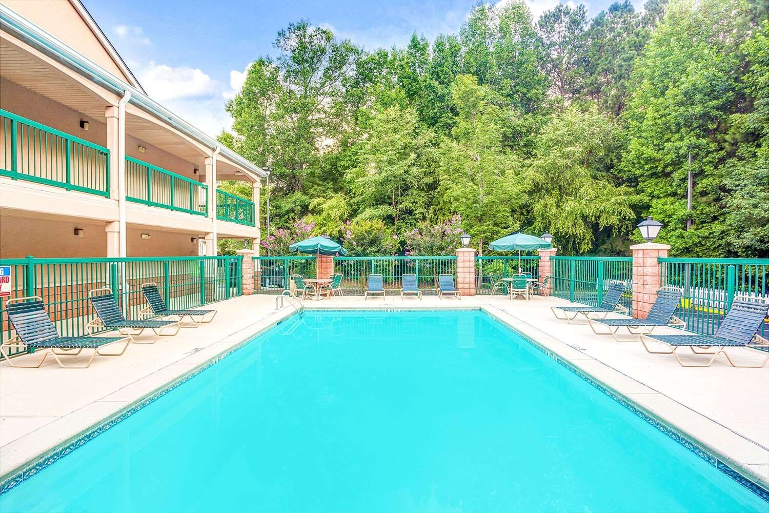 A swimming pool with lounge chairs and umbrellas, surrounded by a fence, with a building in the background.