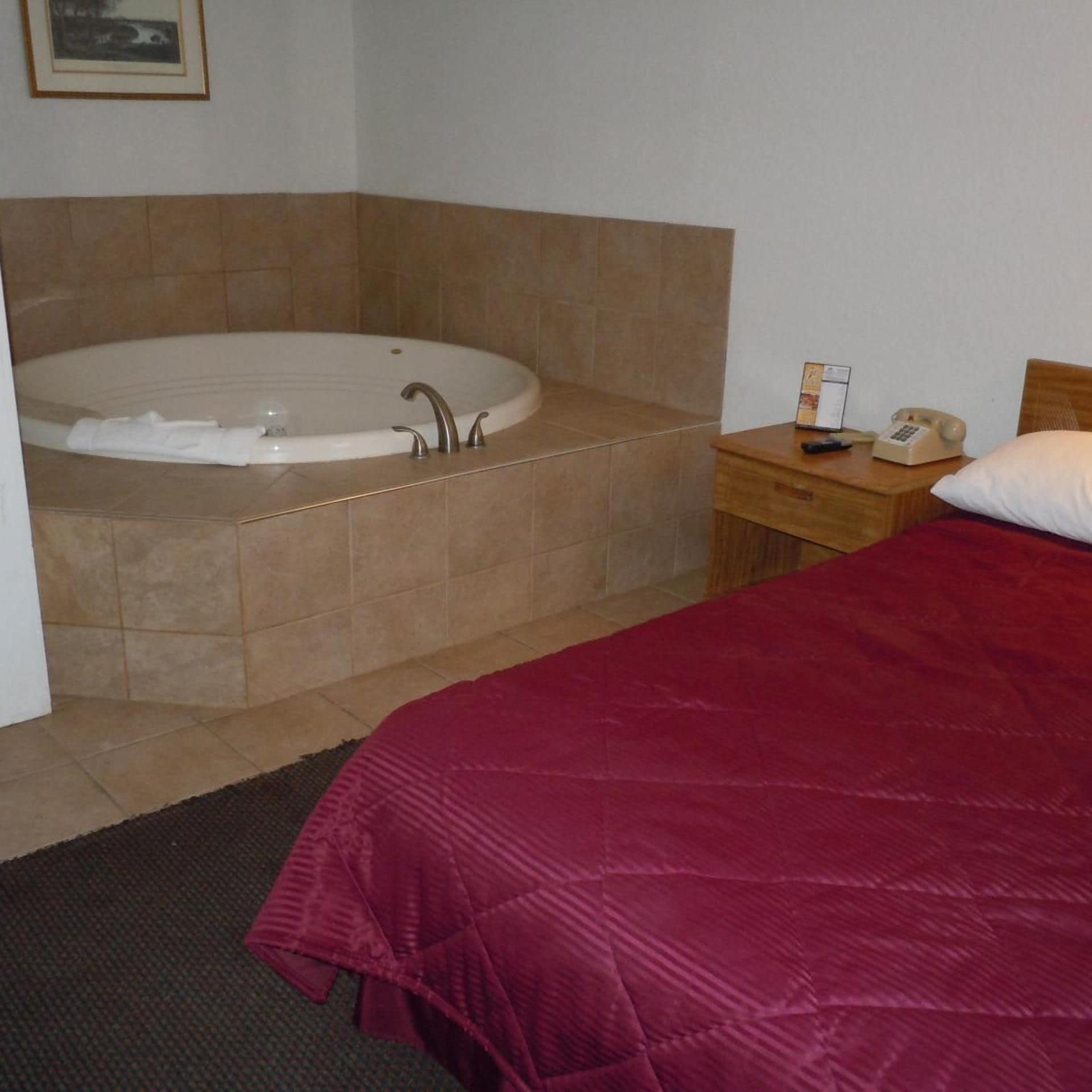 A hotel room with a large bathtub, a wooden nightstand with a telephone, and a bed with a red blanket.