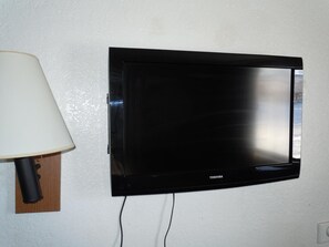 A flat-screen TV mounted on a wall with a lamp on a wooden stand to its left. - Travelodge by Wyndham Grand Rapids North (Grand Rapids)