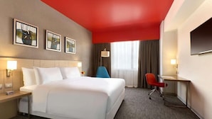 A hotel room with a large bed, a desk, a chair, and artwork on the wall.