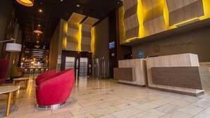 A modern hotel lobby with a reception desk, red seating, and a large screen.