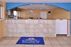Un área de recepción con mostrador, una puerta con la leyenda 'Solo personal' y un felpudo azul con el nombre y la ubicación del hotel.