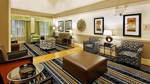 A spacious living room with a large sofa, a patterned ottoman, and a coffee table. There are two armchairs, a side table with a lamp, and a large decorative wall piece. - Country Inn & Suites By Radisson Evansville IN (Evansville)