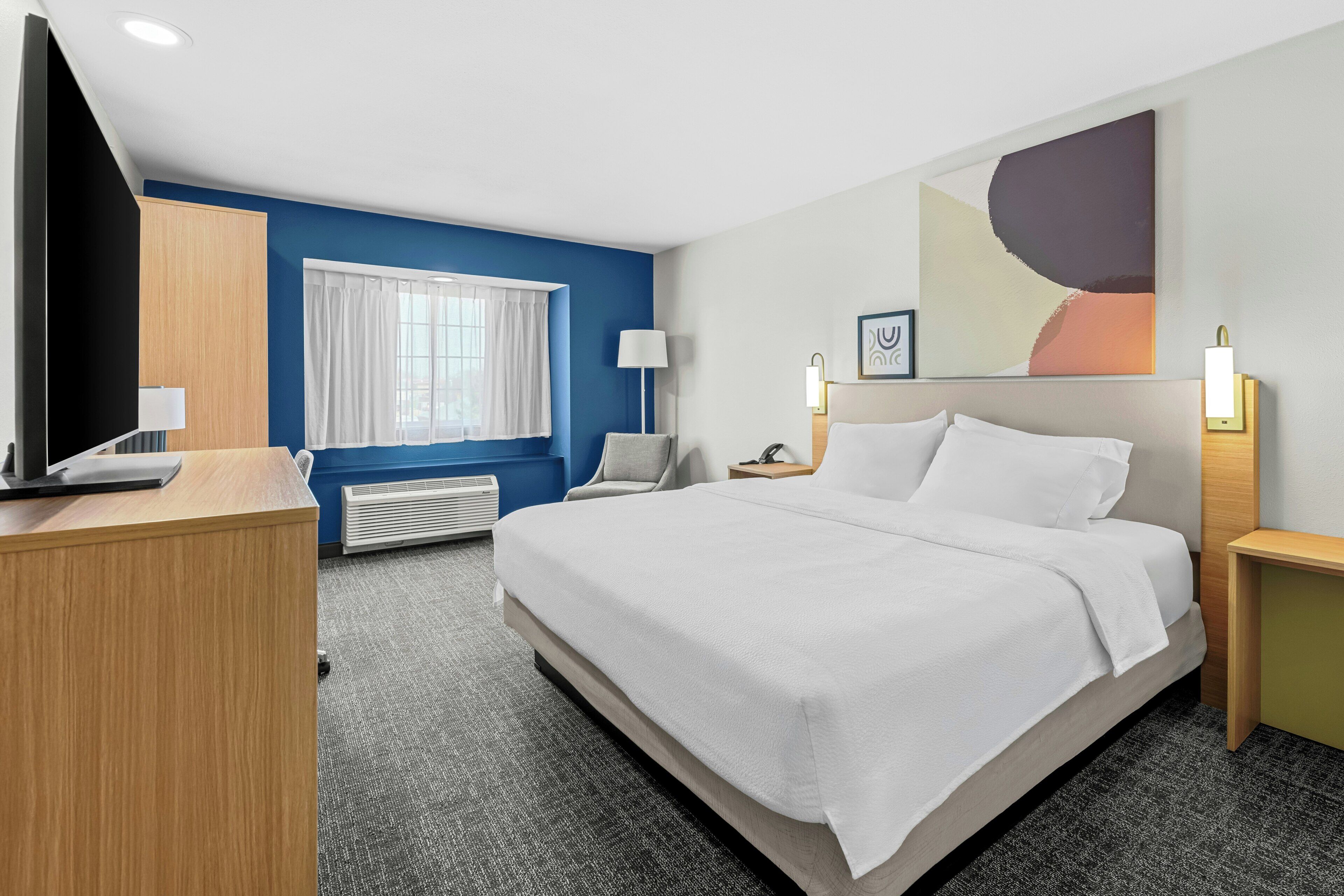 A hotel room with a large bed, a desk, a television, a window with curtains, a wall-mounted lamp, and a painting.