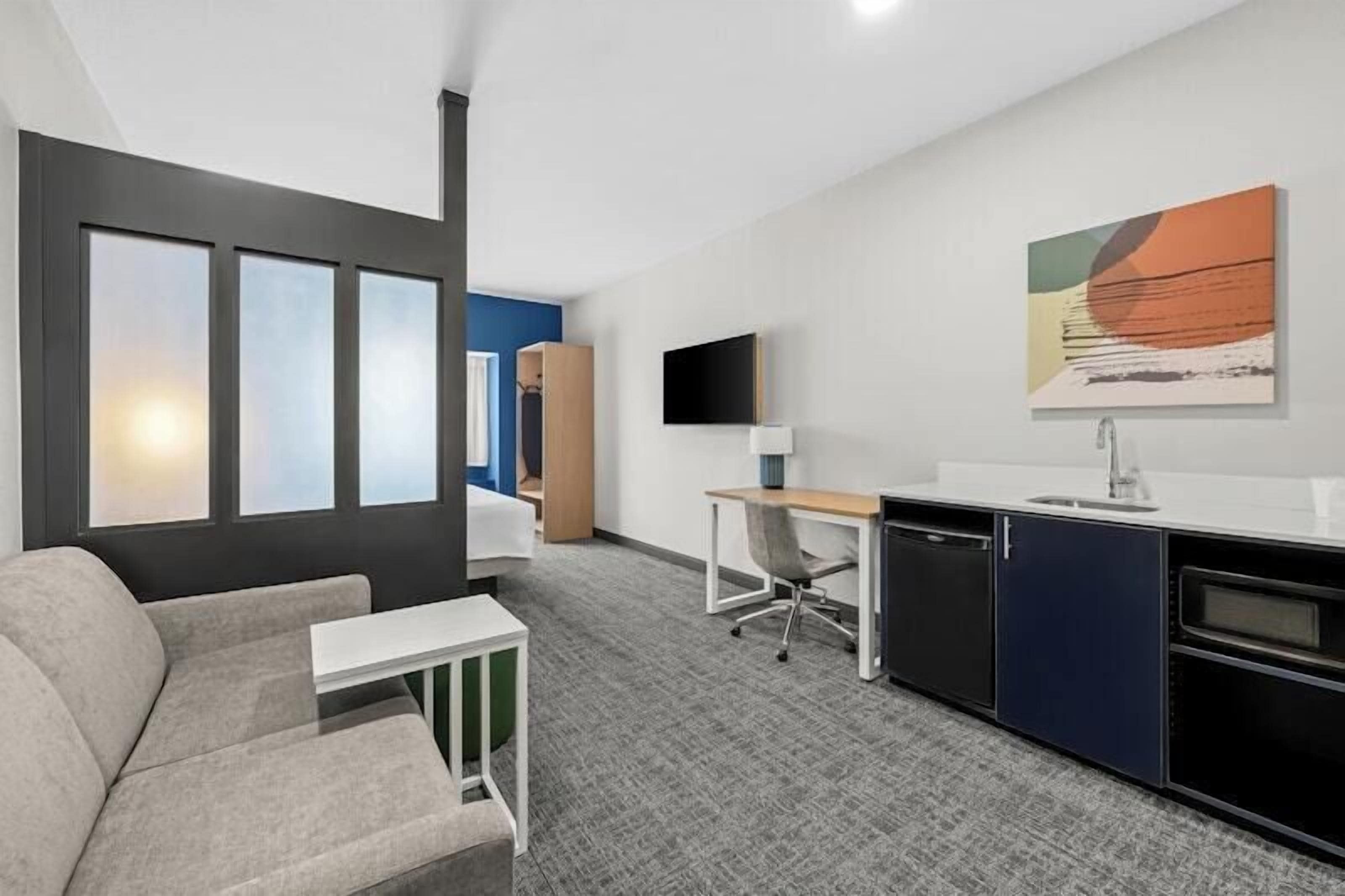 A modern hotel room with a sofa, a desk, a chair, a TV, and a kitchen area.