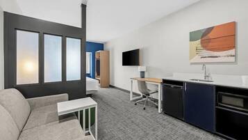 A modern hotel room with a sofa, a desk, a chair, a TV, and a kitchen area.