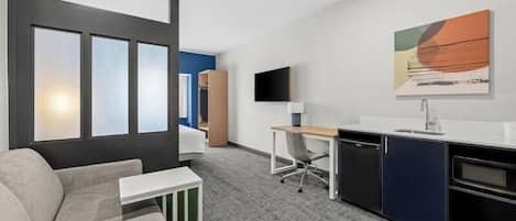 A modern hotel room with a sofa, a desk, a chair, a TV, and a kitchen area.