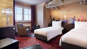 Quarto de hotel com duas camas, uma escrivaninha, uma cadeira, um abajur, um radiador e uma janela.