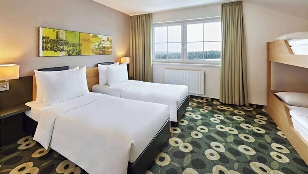 A hotel room with two beds, a window with curtains, a radiator, and a patterned carpet. - Lindner Hotel Nurburgring Motorsport, Part of JDV by Hyatt (Nuerburg)