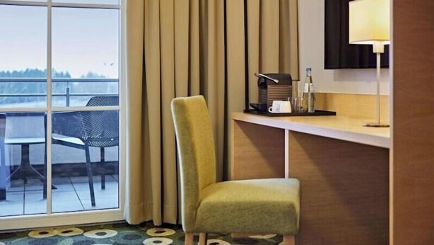 Quarto de hotel com uma janela grande, uma cadeira, uma escrivaninha com luminária e vista para o exterior.