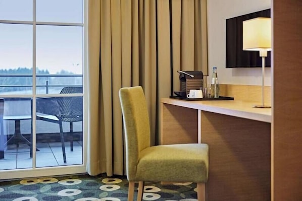 A hotel room with a large window, a chair, a desk with a lamp, and a view of the outdoors. - Lindner Hotel Nurburgring Motorsport, Part of JDV by Hyatt (Nuerburg)