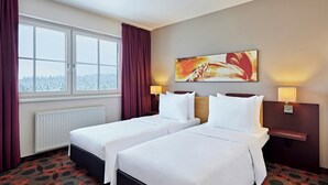 A hotel room with two beds, a window with curtains, a radiator, a painting on the wall, and two bedside lamps. - Lindner Hotel Nurburgring Motorsport, Part of JDV by Hyatt (Nuerburg)