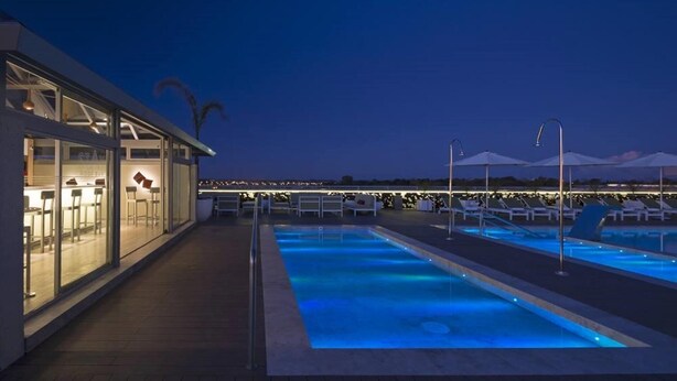 An outdoor swimming pool area with lounge chairs and umbrellas, a bar area with seating, and a view of the city skyline at night.