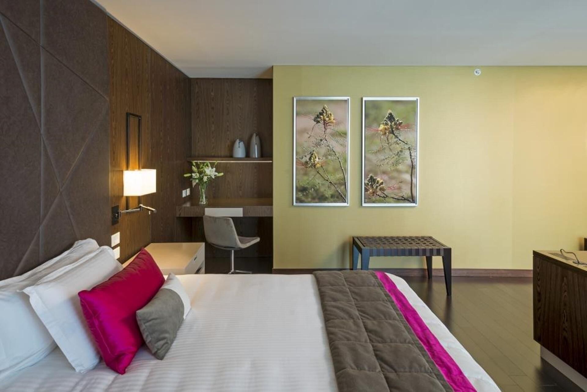 Quarto de hotel com uma cama, uma escrivaninha, uma cadeira e duas fotografias emolduradas na parede.