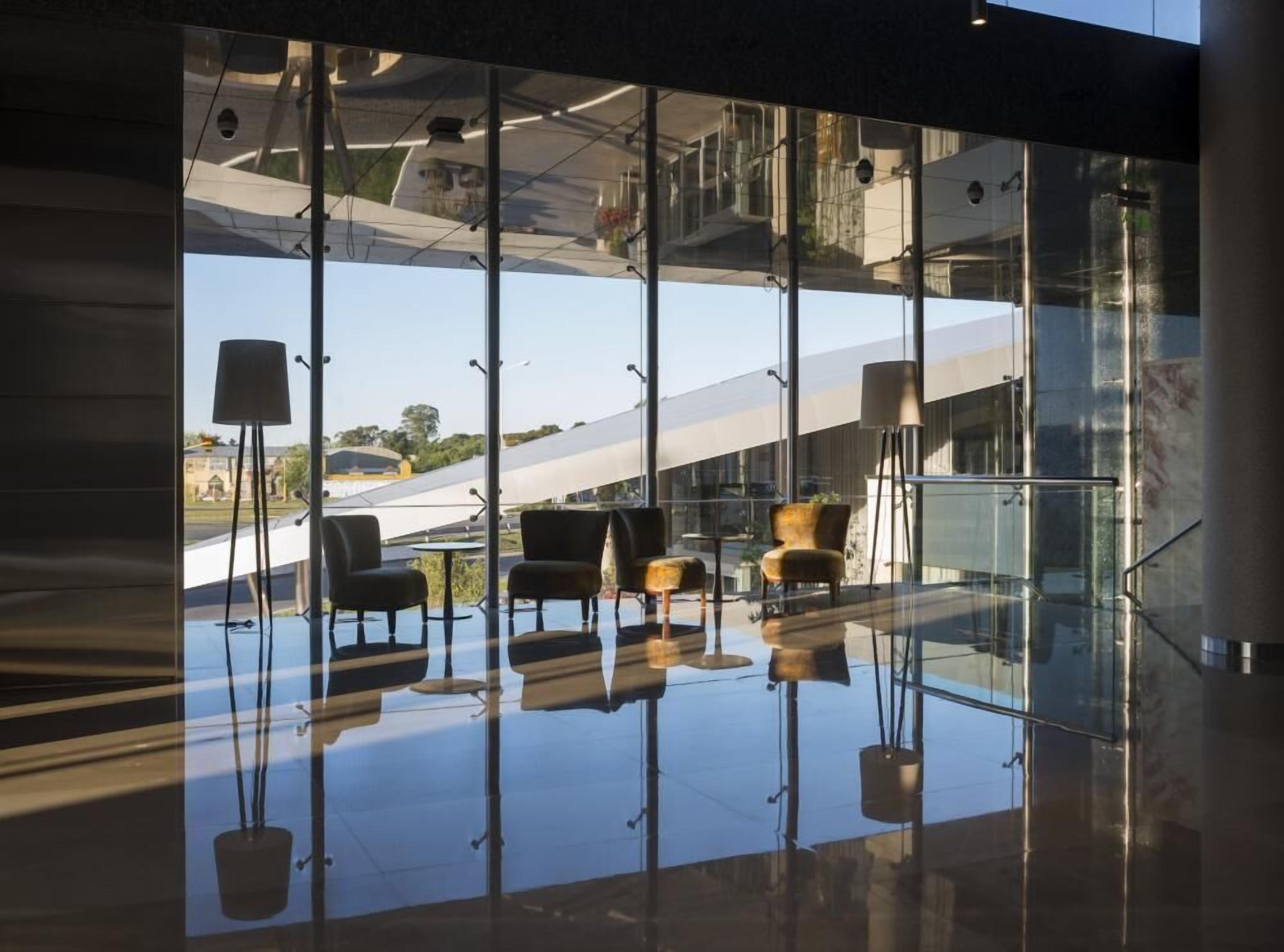 Um interior moderno com parede de vidro, piso refletivo e área de estar com cadeiras e mesa.