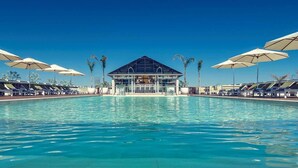 Una piscina con sillones de descanso y sombrillas, un edificio con fachada de vidrio y palmeras.
