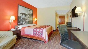 Habitación de hotel con cama, dos lámparas de noche, una silla, un escritorio, un televisor y un cuadro grande en la pared.