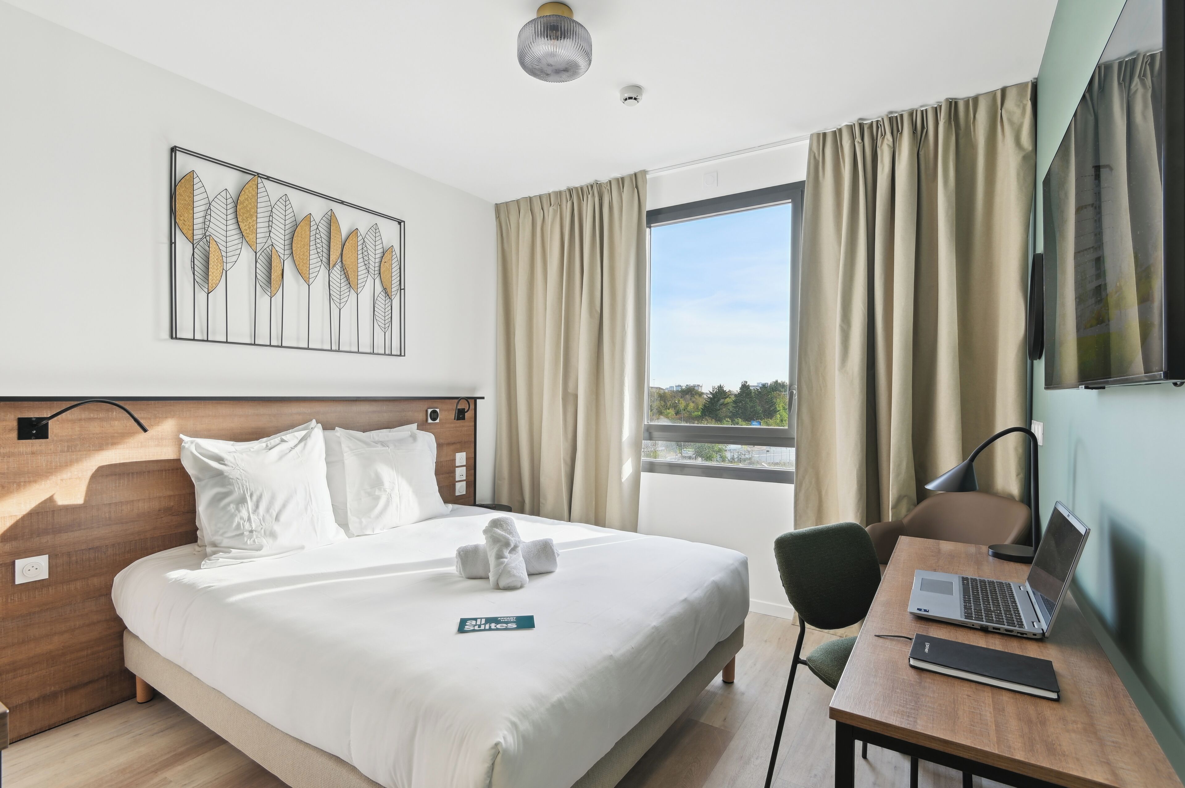 A hotel room with a bed, a desk with a laptop, a chair, a TV, and a window with curtains.