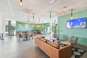 A modern interior with a green wall, a large TV screen, and a mix of seating arrangements including a leather sofa and round tables. - All Suites Appart Hotel Paris Ouest Colombes (Colombes)