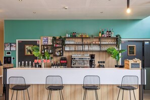 Un espace bar comprenant un comptoir, des tabourets de bar, des étagères remplies de bouteilles et de verres, ainsi qu’une machine à café.