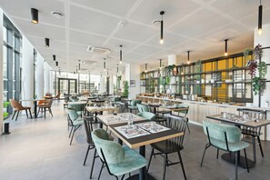 A modern restaurant interior with high ceilings, pendant lighting, and a central serving area. - All Suites Appart Hotel Paris Ouest Colombes (Colombes)
