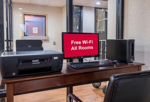 A hotel room with a desk featuring a printer, a monitor, and a computer. A sign indicates free Wi-Fi is available in all rooms. - Wingate by Wyndham Augusta I-20 (Augusta)