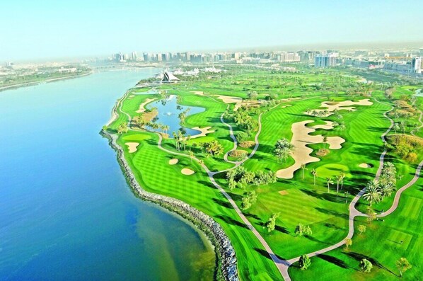 Un campo de golf con un cuerpo de agua, silueta urbana y edificios modernos al fondo.