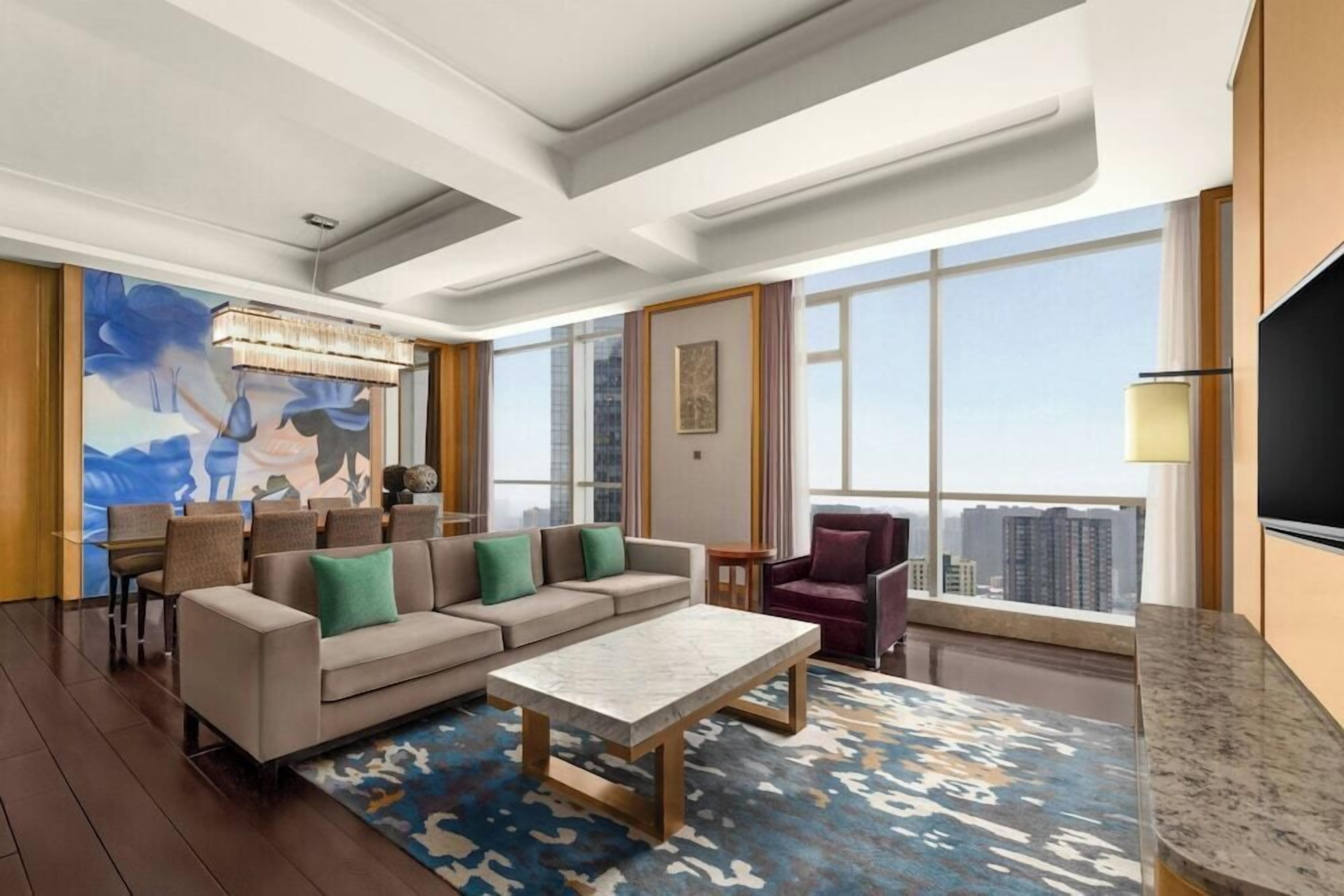 A modern living room with a sofa, coffee table, and chairs, large windows with a city view, and a mounted TV.