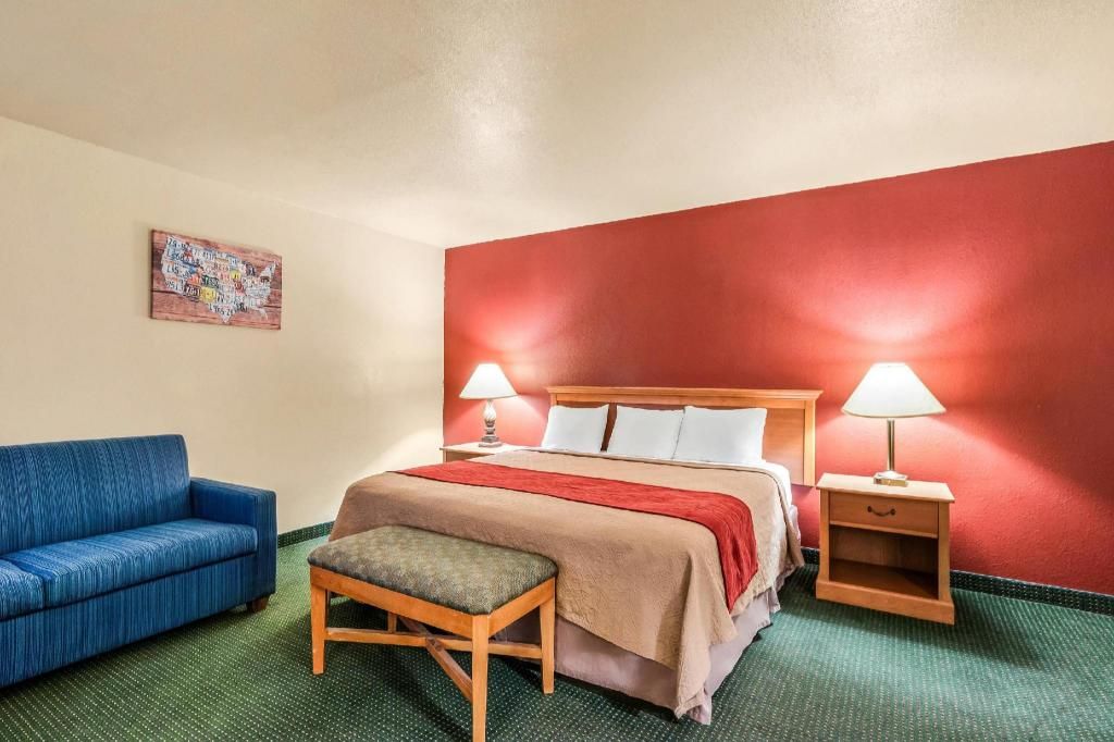 A hotel room with a bed, two bedside tables with lamps, a blue sofa, a wooden bench, and a red accent wall.