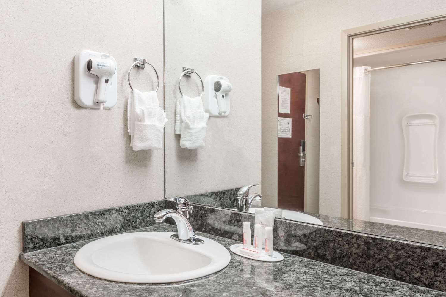 Une salle de bain dotée d’un comptoir en granit, d’un lavabo, d’un miroir et de deux sèche-cheveux fixés au mur.